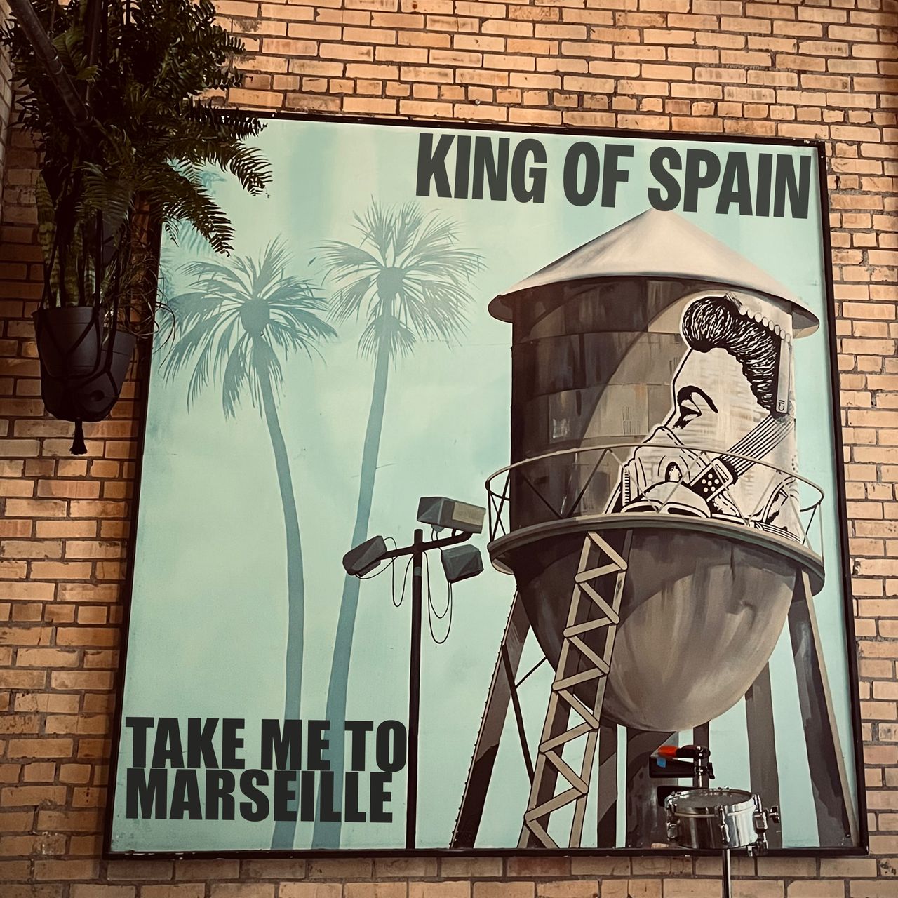 Single artwork for Take Me To Marseille by King of Spain, featuring palm trees and an illustrated water tower poster mounted on a brick wall.